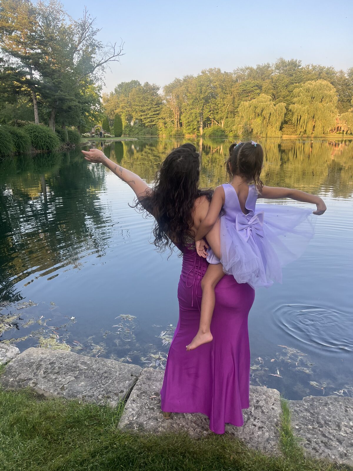 Adelina holding her daughter at the edge of a lake, both reaching toward the water